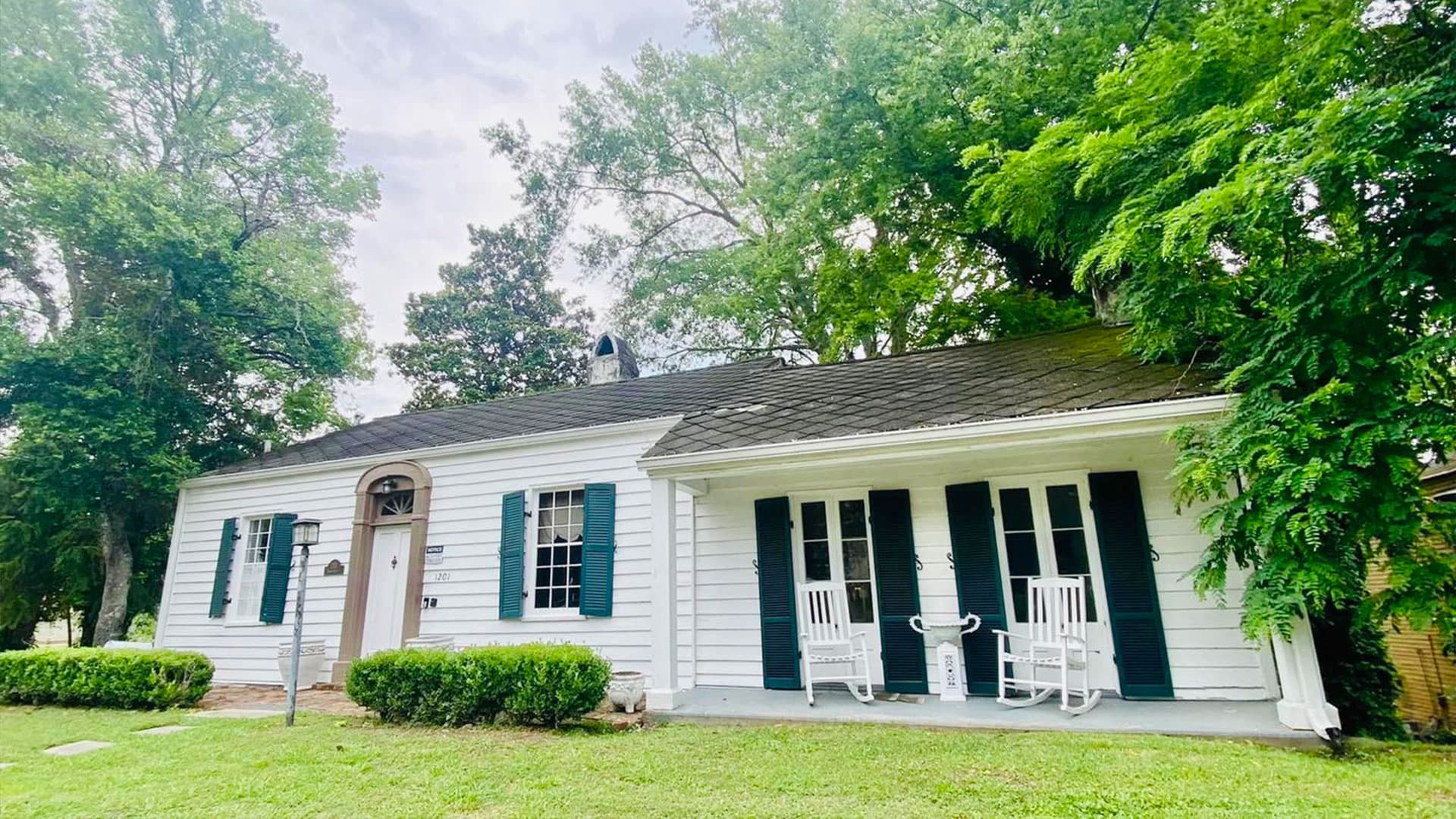 A charming white cottage with black shutters and rocking chairs on the porch, surrounded by green trees and grass.